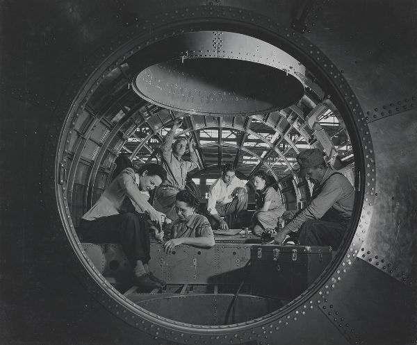 Framed through a circular opening, three men and three women work together inside a partially finished airplane chassis.