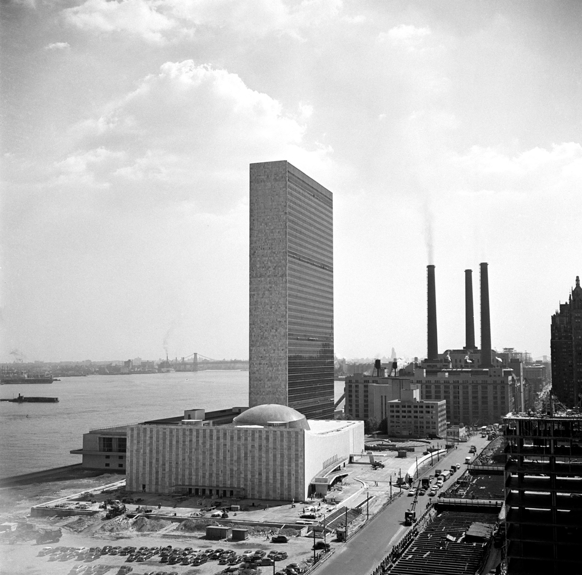 A wide-angle photograph of a low rectangular building in front of a skyscraper on the twentieth-century industrial riverfront of New York City.