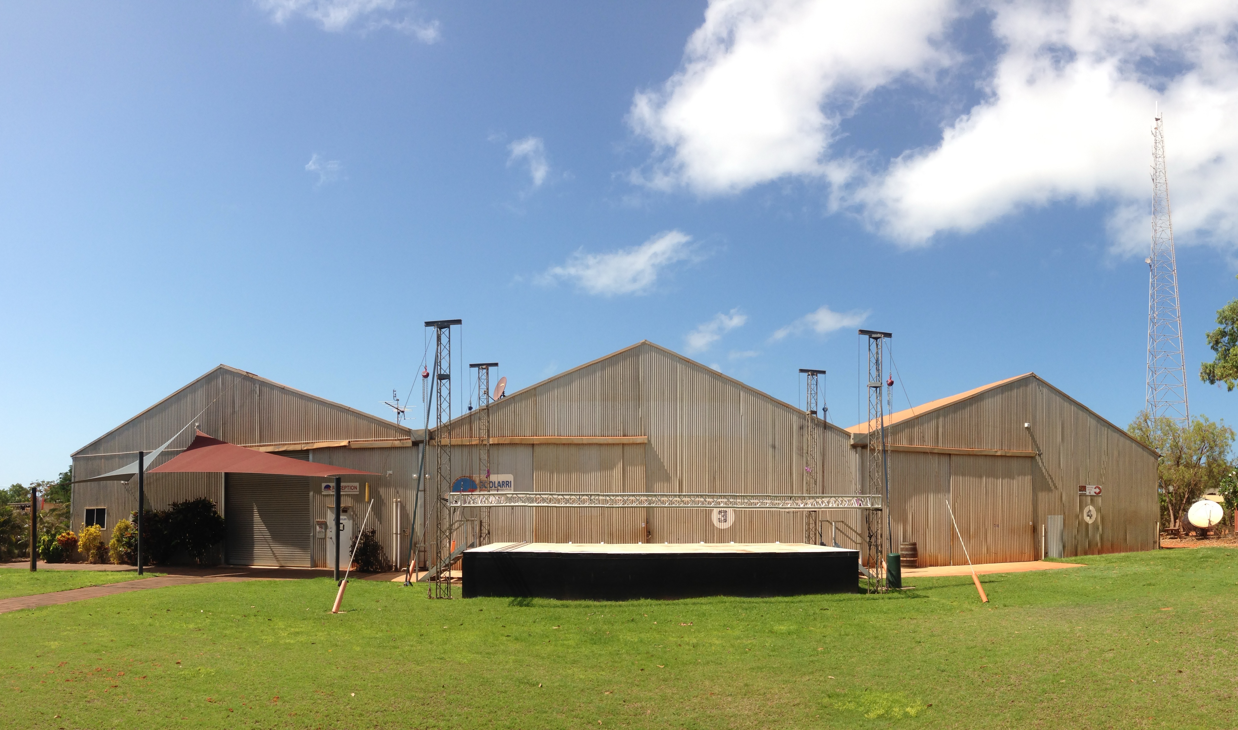 Panoramic view of the three large corrugated iron sheds that include PAKAM and Goolarri. Below them is an outdoor stage surrounded by grass.