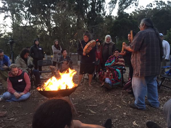 It is twilight and a fire pit glows. Next to it, a woman holds a hand drum and a stick, singing. People with name tags sit and stand around the fire.