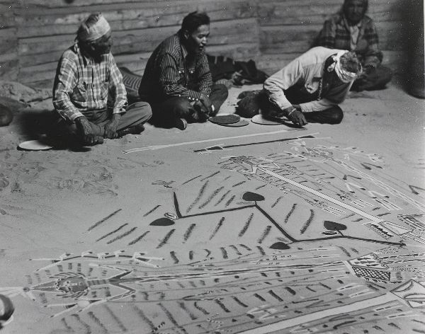 Three men sit at the edge of a ceremonial drawing: the central man, watched by his right-hand neighbor, sings, while the third creates the drawing.