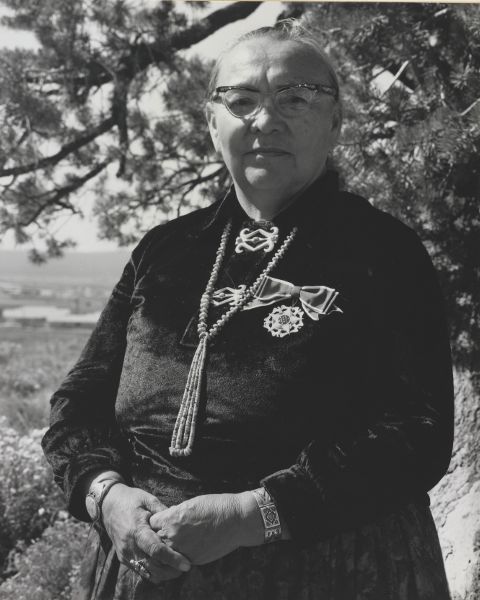Middle-aged woman, in dark velvet blouse, silver and beaded jewelry, and medal on a ribbon bow, looks at the camera with neutral expression.