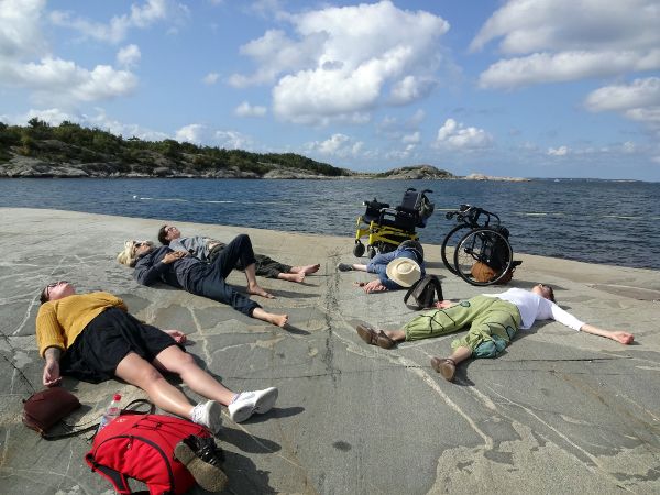 Five people laying on a rock beside a body of water; two wheelchairs sit nearby.
