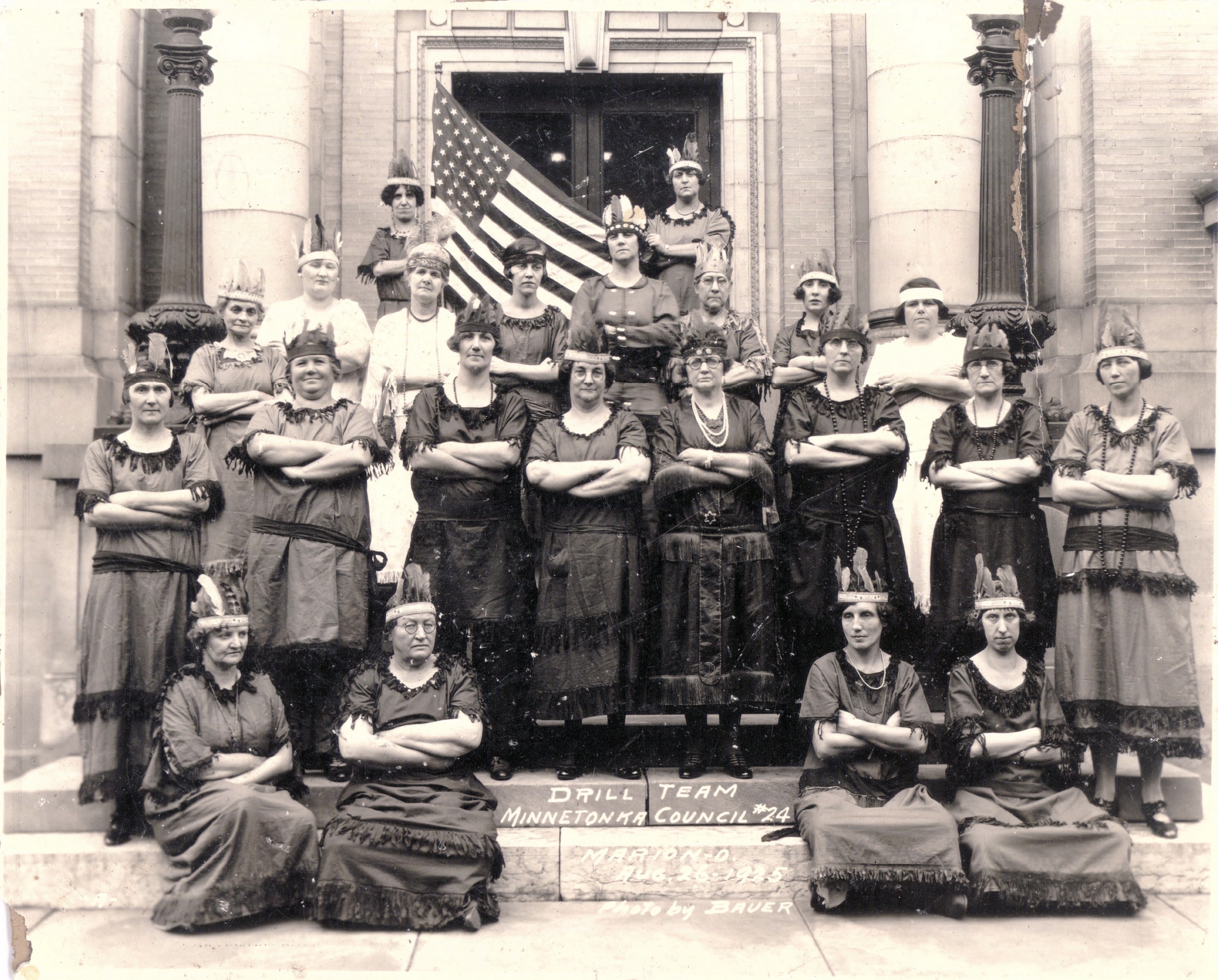 A group of white women are wearing Native American costumes posed with arms crossed in front of a brick building. One holds an American flag.