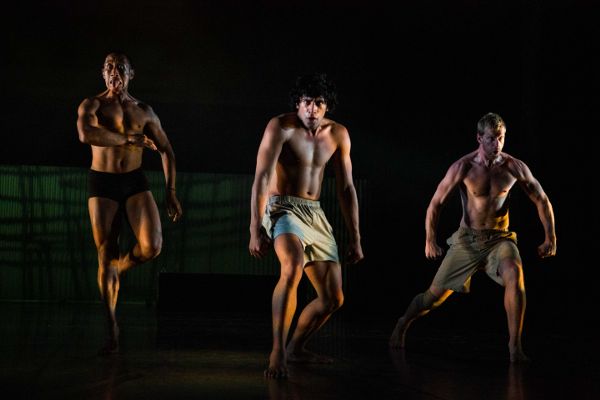 Three male dancers in shorts with defined muscles face front. One jumps, arm bent, tongue out. One clenches fists. One flexes arms down, knee bent.