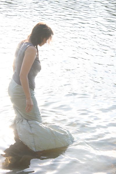 A woman walks into water up to her knees. She is bent slightly forward, looking down. Her arms hang down and her skirt billows out beside her.