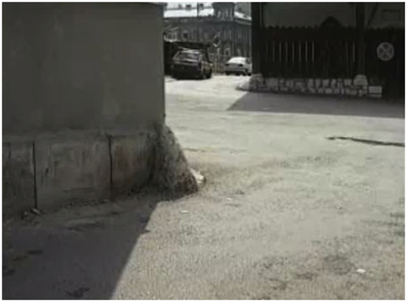 A low-angle image showing the corner of a brown building beside a twisting alleyway with a few par parked cars in the background.