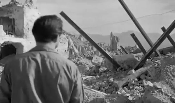 A man with his back to the camera stares at a site of mass architectural destruction and a city in ruins.