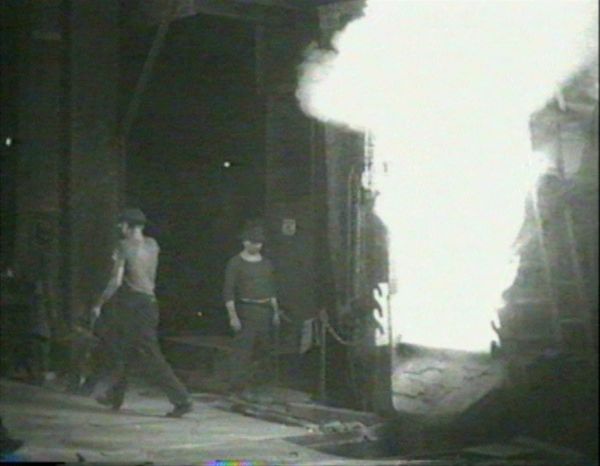 A large fire flares in an industrial building as two men in work clothes casually stand by.