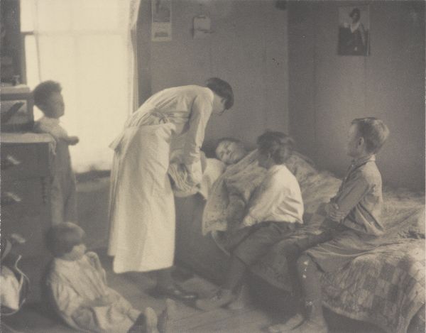 A woman in nurse’s uniform shows a newborn baby to its mother, in bed at home, as four other children look on.
