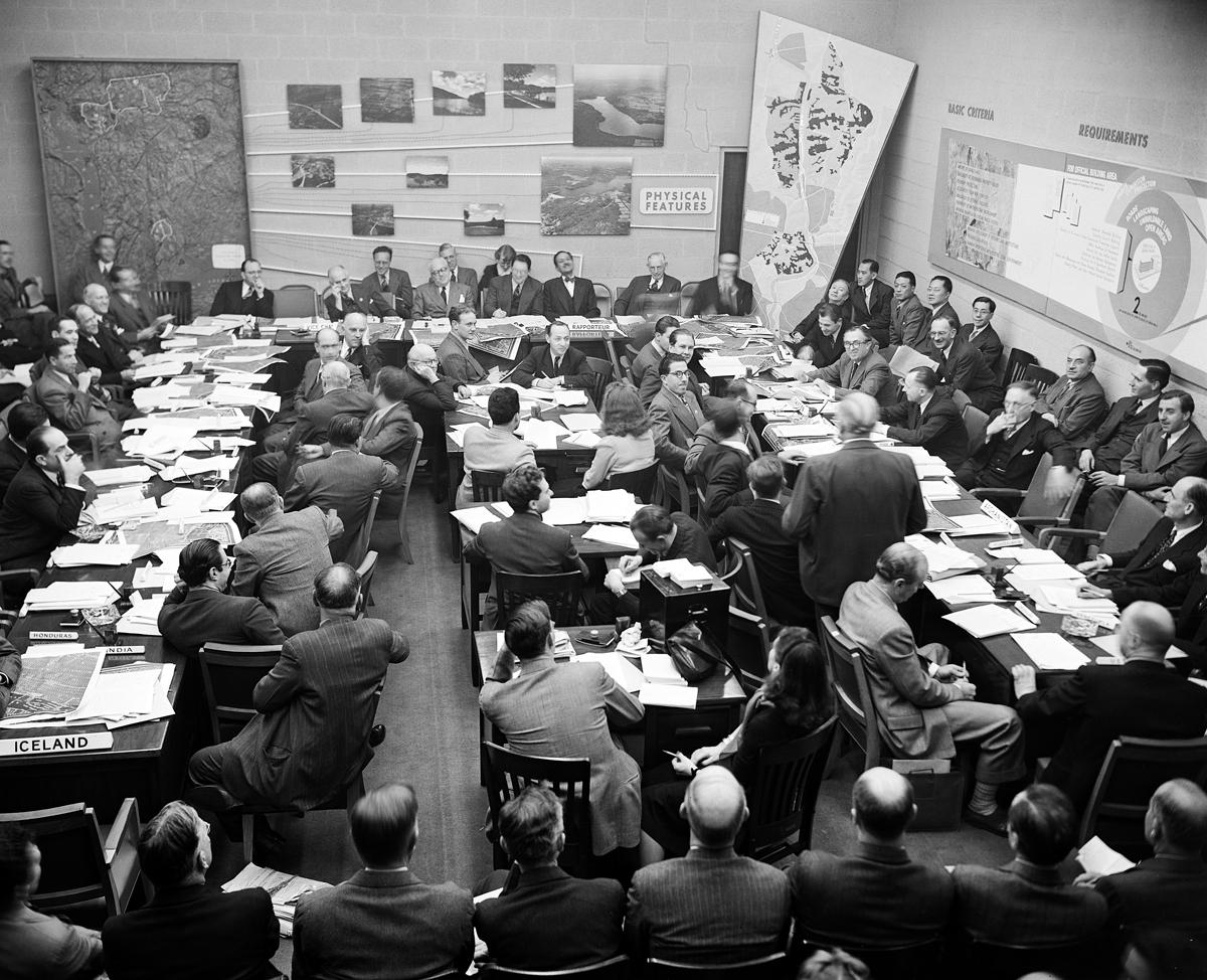 Photograph of room with people gathered around tables filled with papers, showing maps and photographs mounted on the walls.