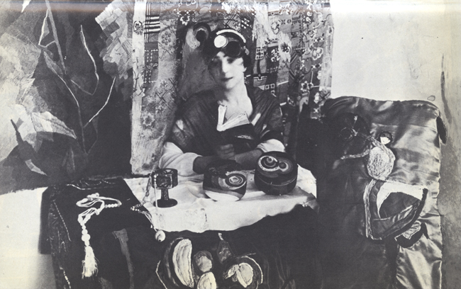 The exhibition features a painting that depicts a woman seated at a table. Behind her, the walls are adorned with paintings, and on the table, various art pieces are arranged. The woman is captured holding a book in her hands.