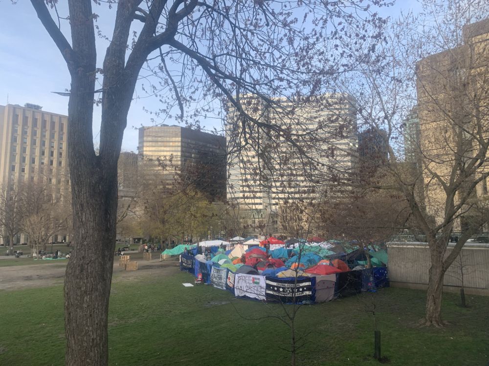 Clustered between two trees, a number of colorful tents sit on the grass at McGill University. Gray buildings are seen in the background as students gather near the tents.