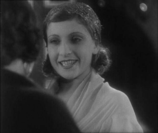 A woman dressed in white speaks with and smiles toward another woman in the foreground, who is out of focus and has her back turned.