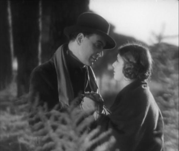 A finely dressed man and a woman wearing a shawl hold hands and stare lovingly into each other’s eyes as they stand in the middle of a field.