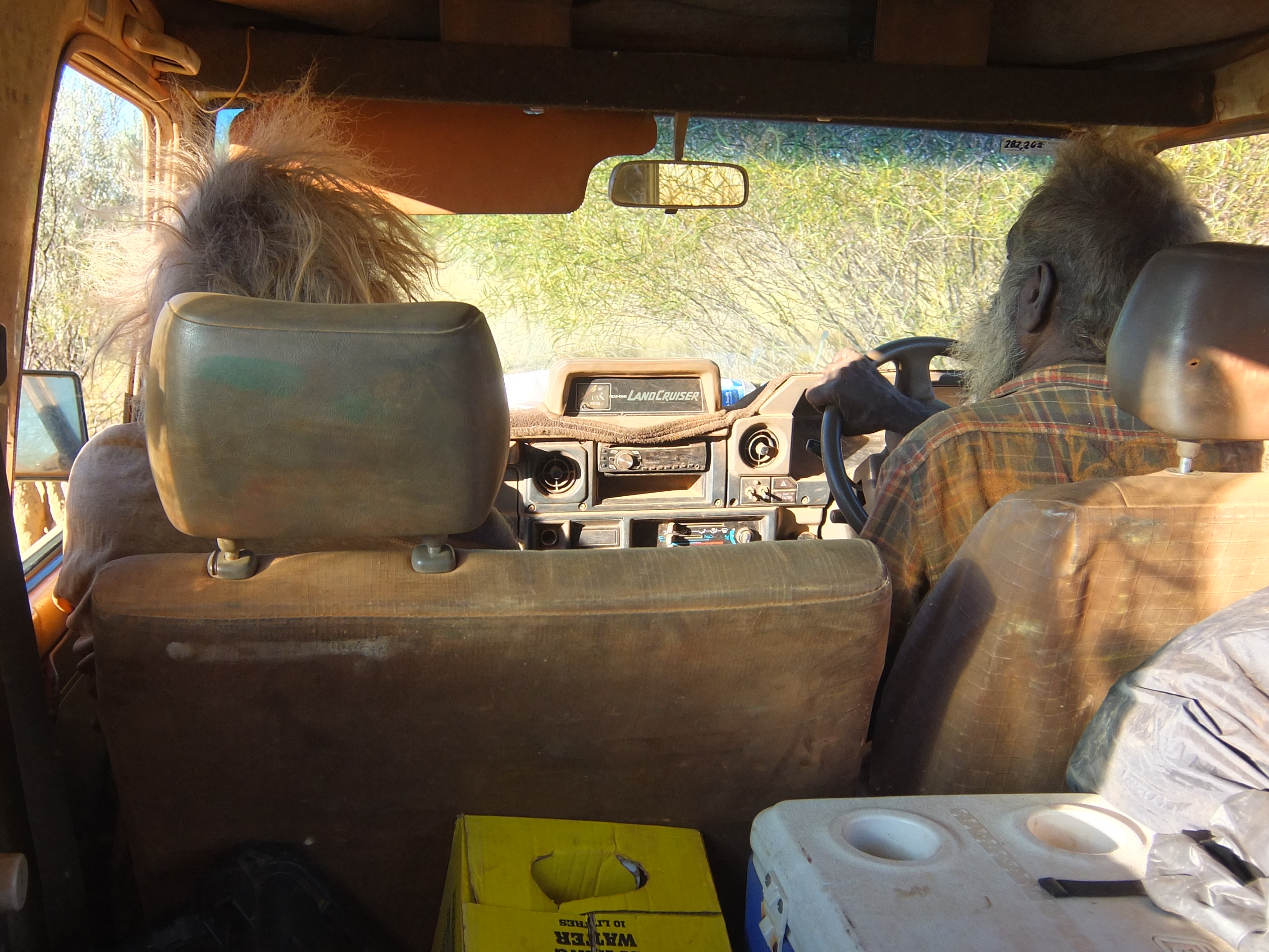 View from the back of a troopy with a cooler and yellow box of water in view. Mark is driving through thin trees, and Larry is in the passenger seat.