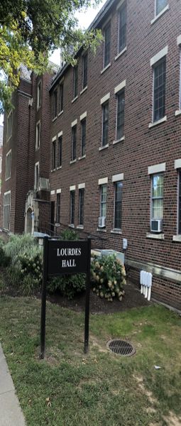 A picture of a brick dormitory building with many windows and an entrance. A sign beside it says “Lourdes Hall.”