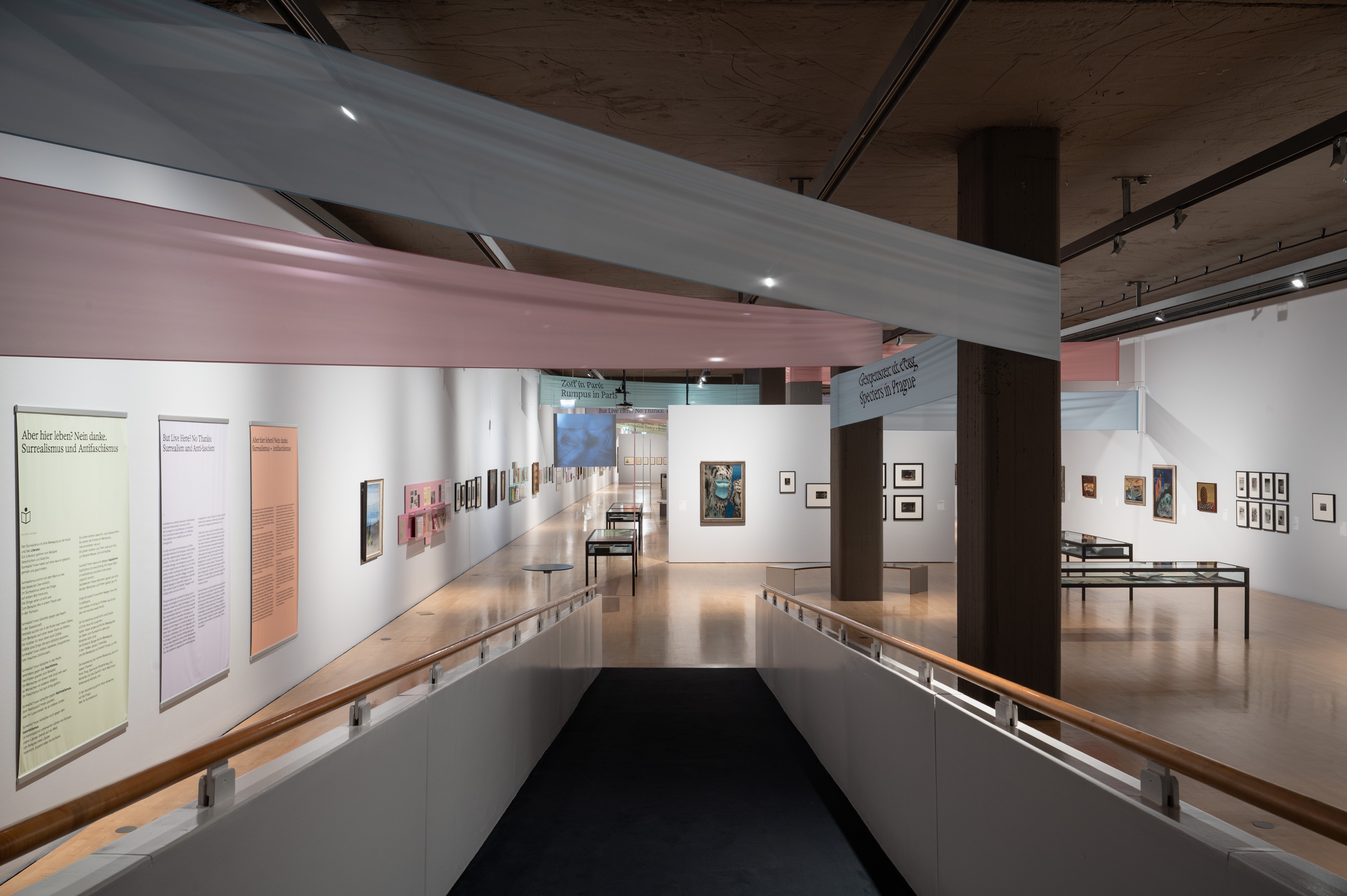 Interior shot of the exhibition space looking down a ramp towards the far back wall. The space is partially bisected by a partition wall protruding from the right-hand side. Art works appear on all sides of the walls and in vitrines.