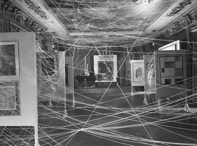 The cover depicts a room adorned with paintings on vertically standing boards. Strings traverse the room, particularly near the ceiling, creating an appearance reminiscent of spider webs.