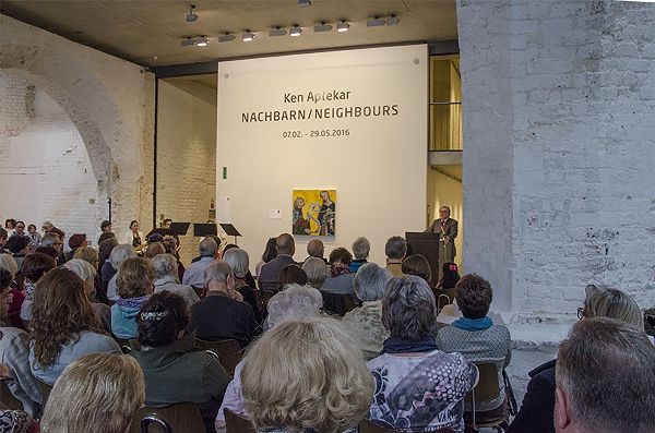 Man at a podium in the background addresses a seated audience in a white brick room.