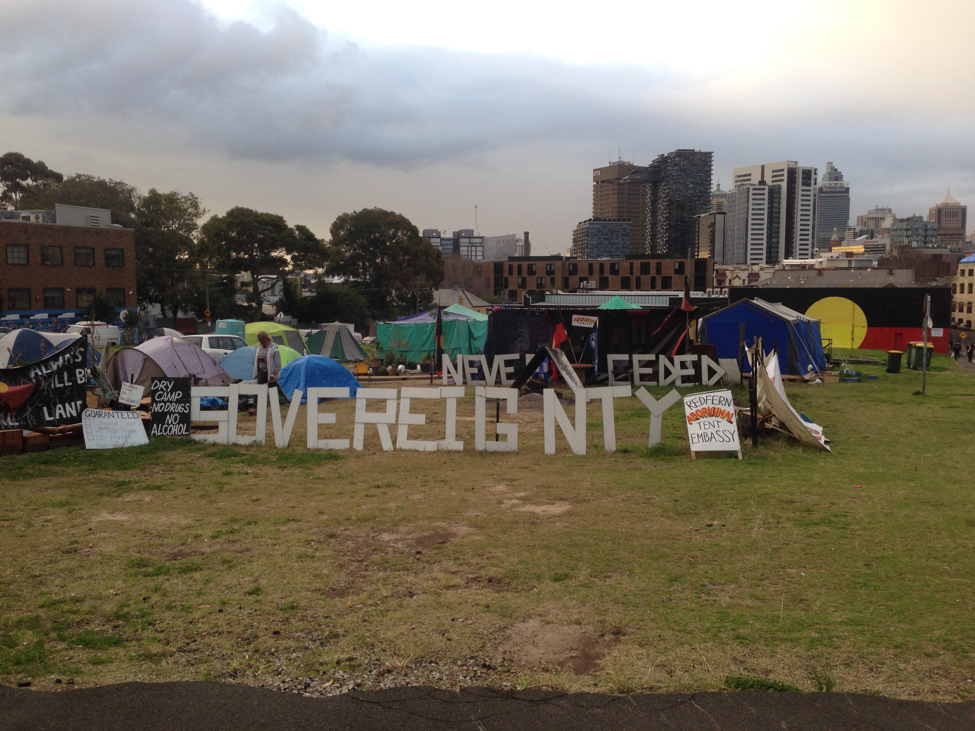 Large white letters spelling Sovereignty Never Ceded sit near dozens of tents. Sydney skyline and a wall painted with Aboriginal flags behind.