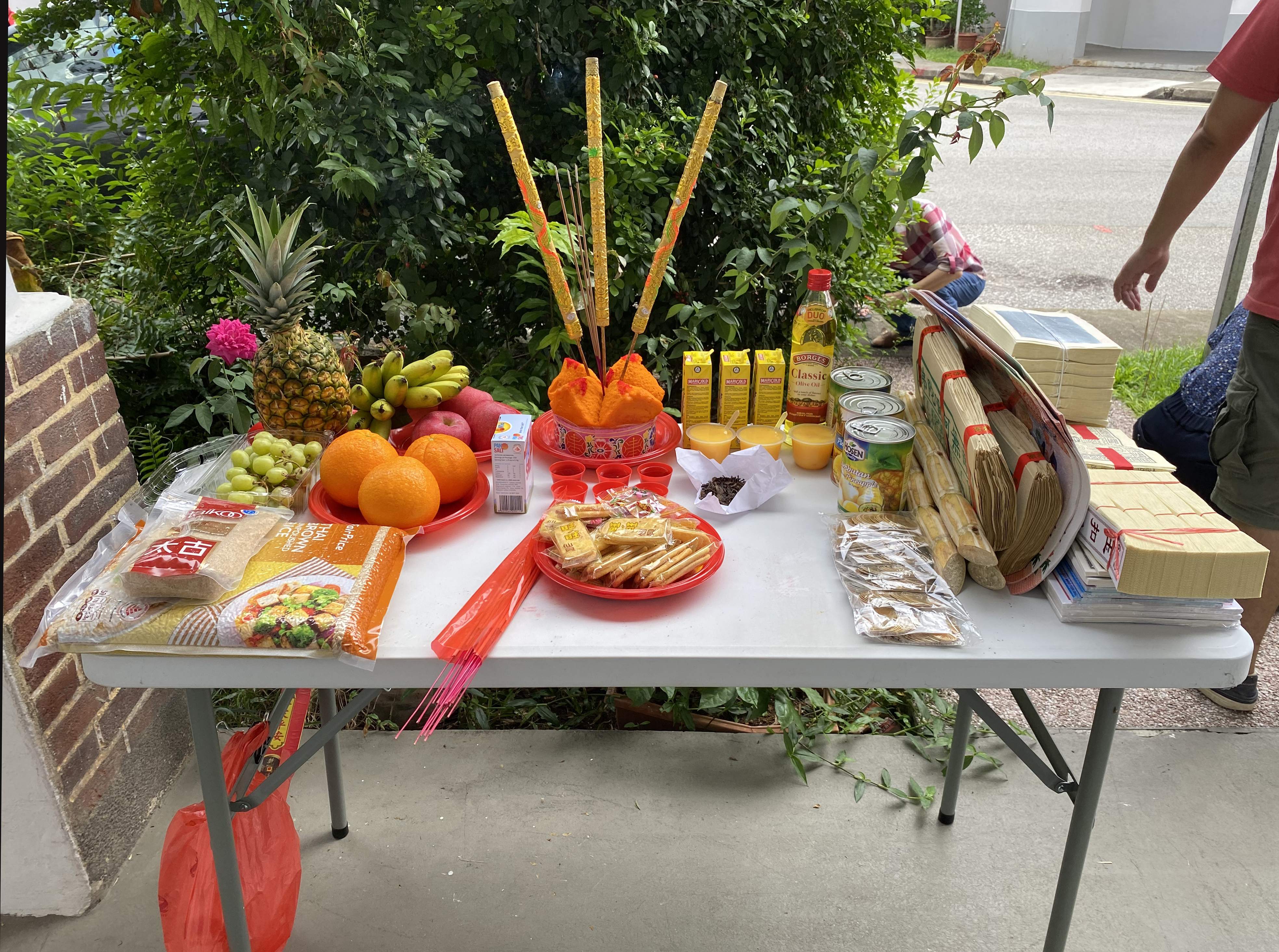 A folding table is covered with plates of fruit, rice, and snacks. Joss paper money is piled to one side, and ceremonial candles are at center.