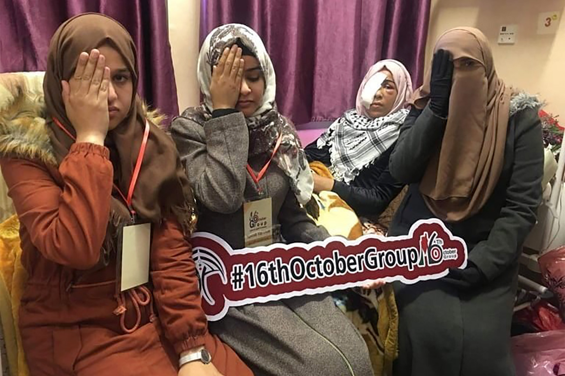 Four women in a hospital room, three of whom cover their right eye with their hands. The fourth woman has a bandaged over her lost eye. They hold a sign reading “16th Otobober Group.”