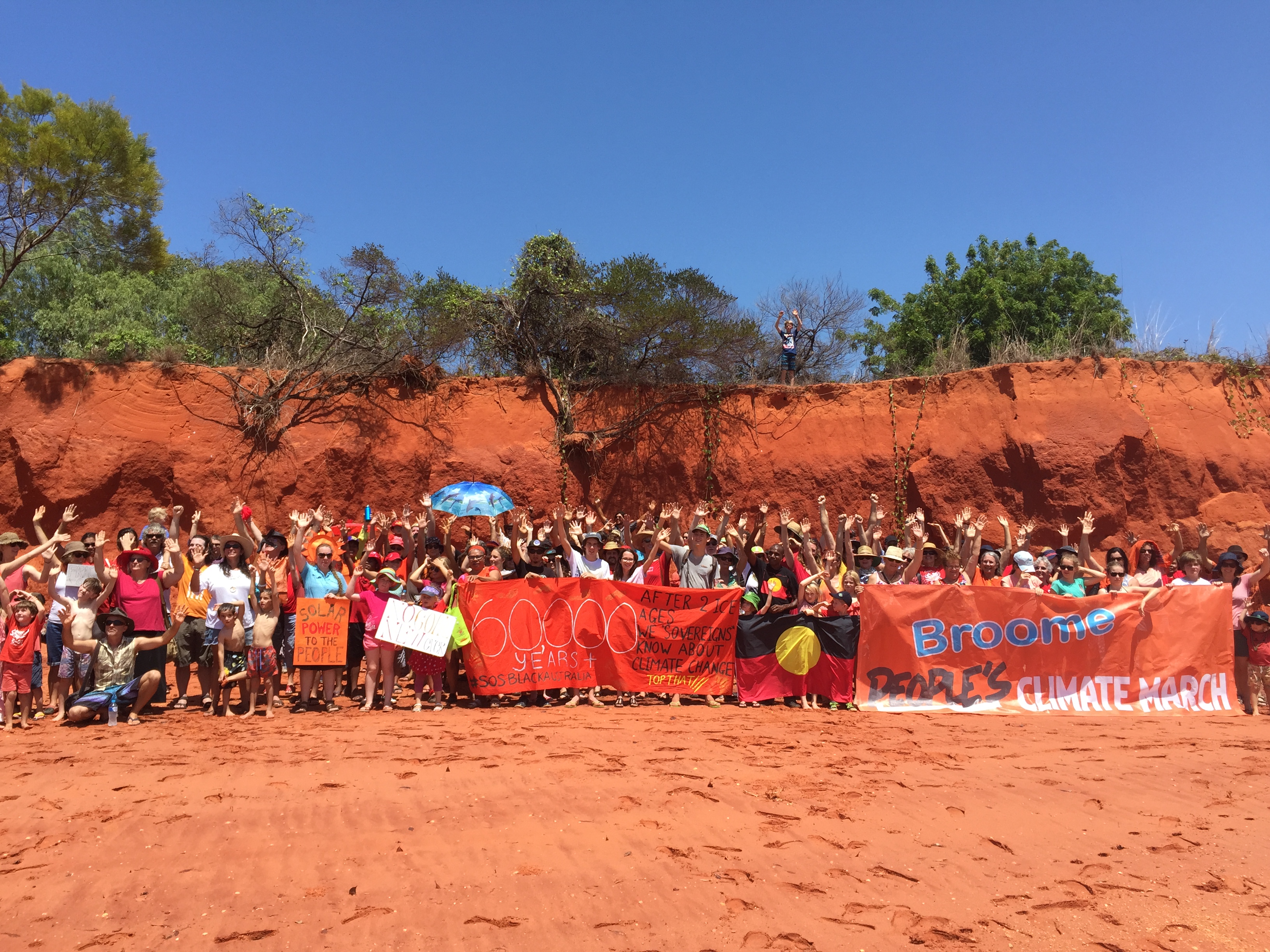 Over a hundred people hold signs and raise their hands at the top of a beach near the bottom of a steep pindan cliff under a sunny sky.
