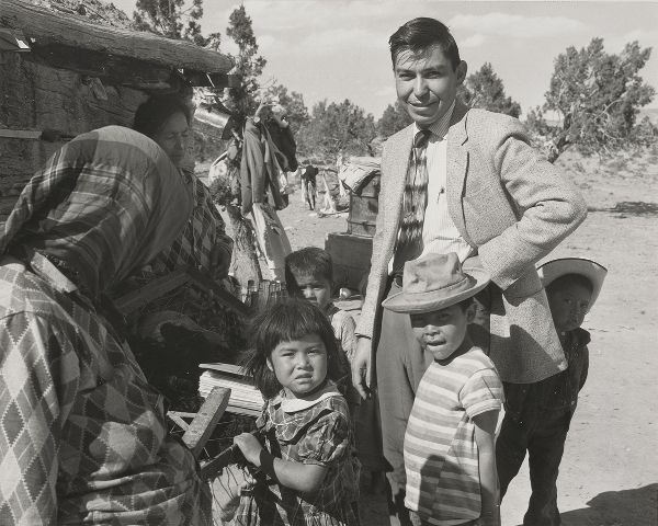 Two women, one man, and four children stand near a hogan in a rural landscape, the man smiling at the camera.