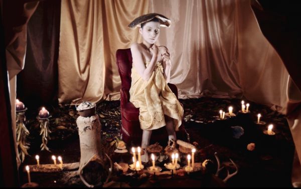Seated figure surrounded by candles and curtains.