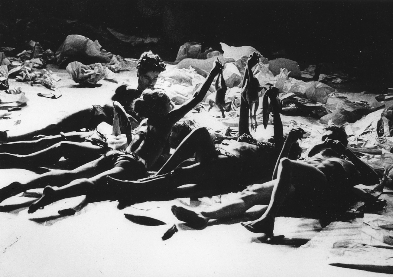 A group of people, James Tenney, Dorothea Rockburne, Carolee Schneemann, Sandra Chew, and Stanley Gochenouer, lie on the ground interacting with objects in a dimly lit space.