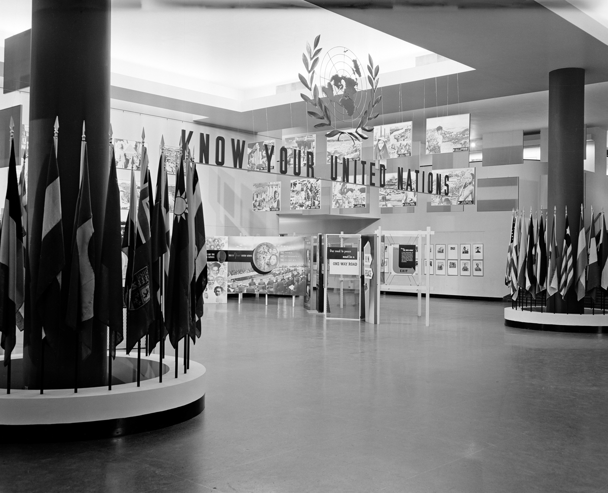 Photo of entrance to an exhibition room with the phrase “Know Your United Nations” hanging from the ceiling between two columns surrounded by flags.