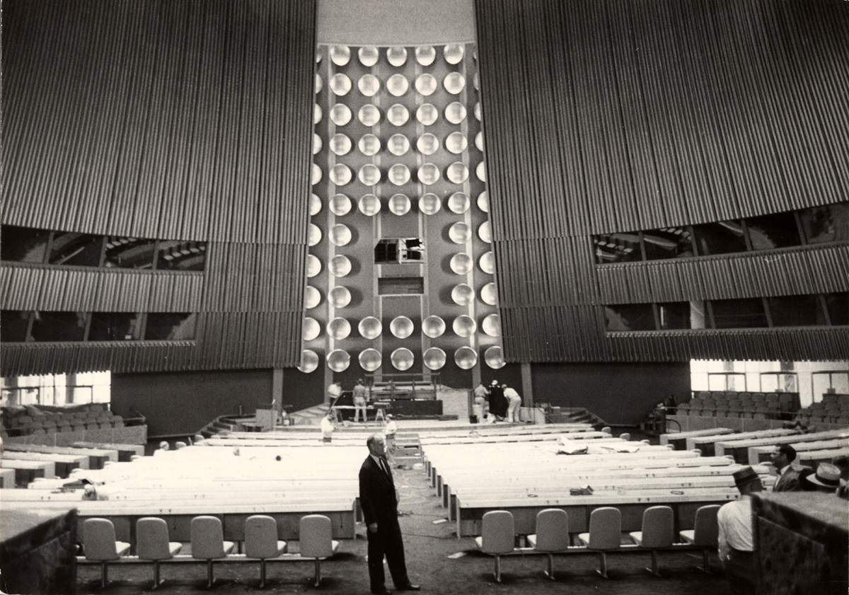 Photograph of man standing in front of an empty plenary hall under construction before the UN Symbol was installed to cover loudspeakers behind.