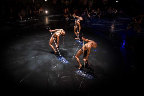 Three male dancers wearing short black shorts, holding flat mops on their right, arched back, looking up. Viewers sit on black floor in background.