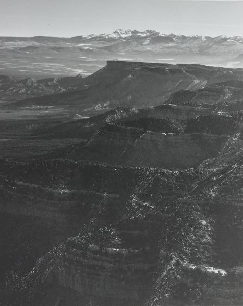 An aerial view of a dark mountain range in the foreground and a mesa in front of snow-capped mountains on the horizon.