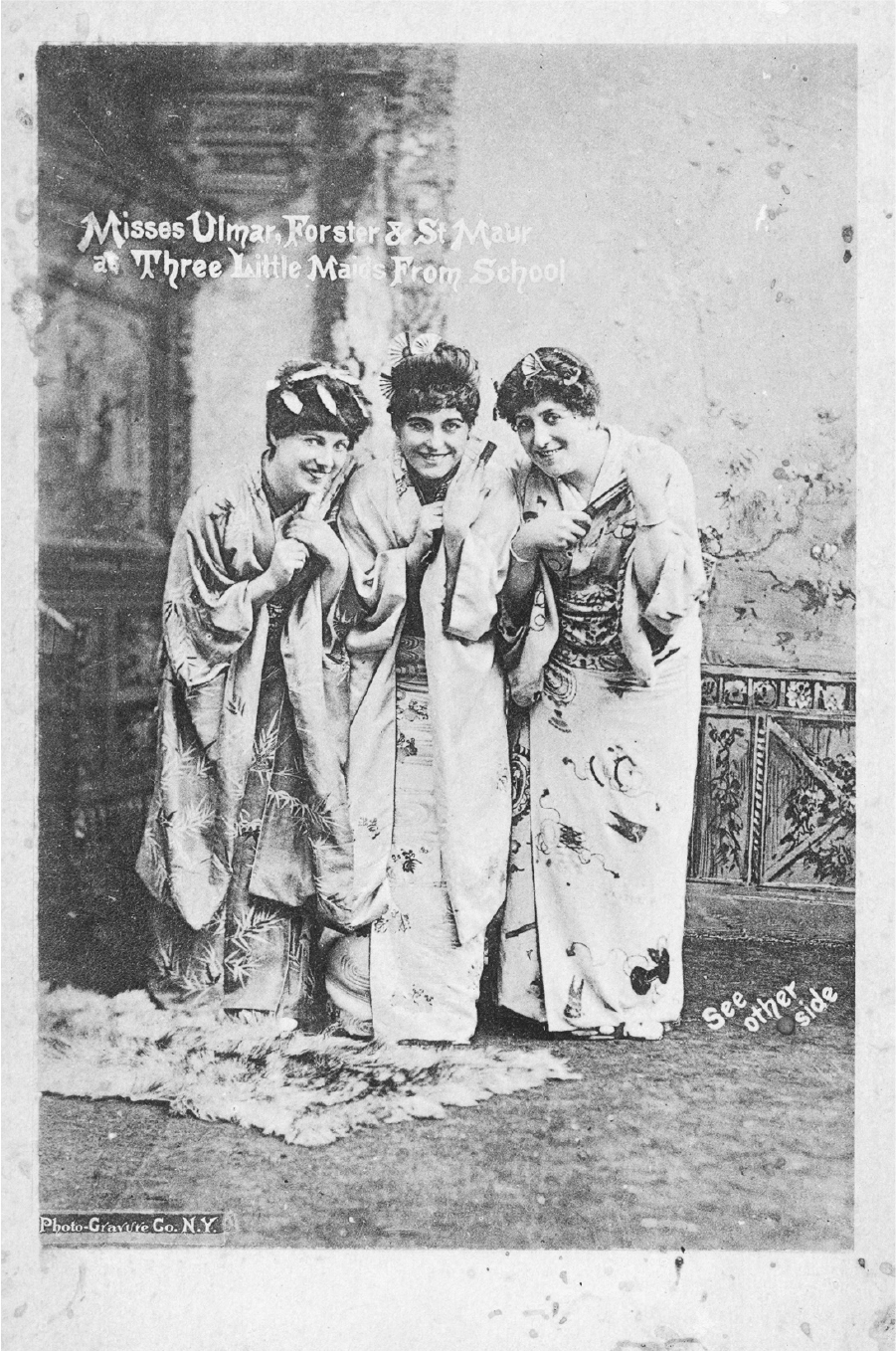 A photograph of the Three Little Maids in traditional attire standing together besides each other with their postures bent a little. They pose with a happy smile on their face.