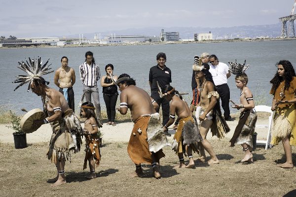Outside before the bay, seven young dancers in leather skirts, two with feather roaches, bend forward, knees lifting. A few people stand watching.