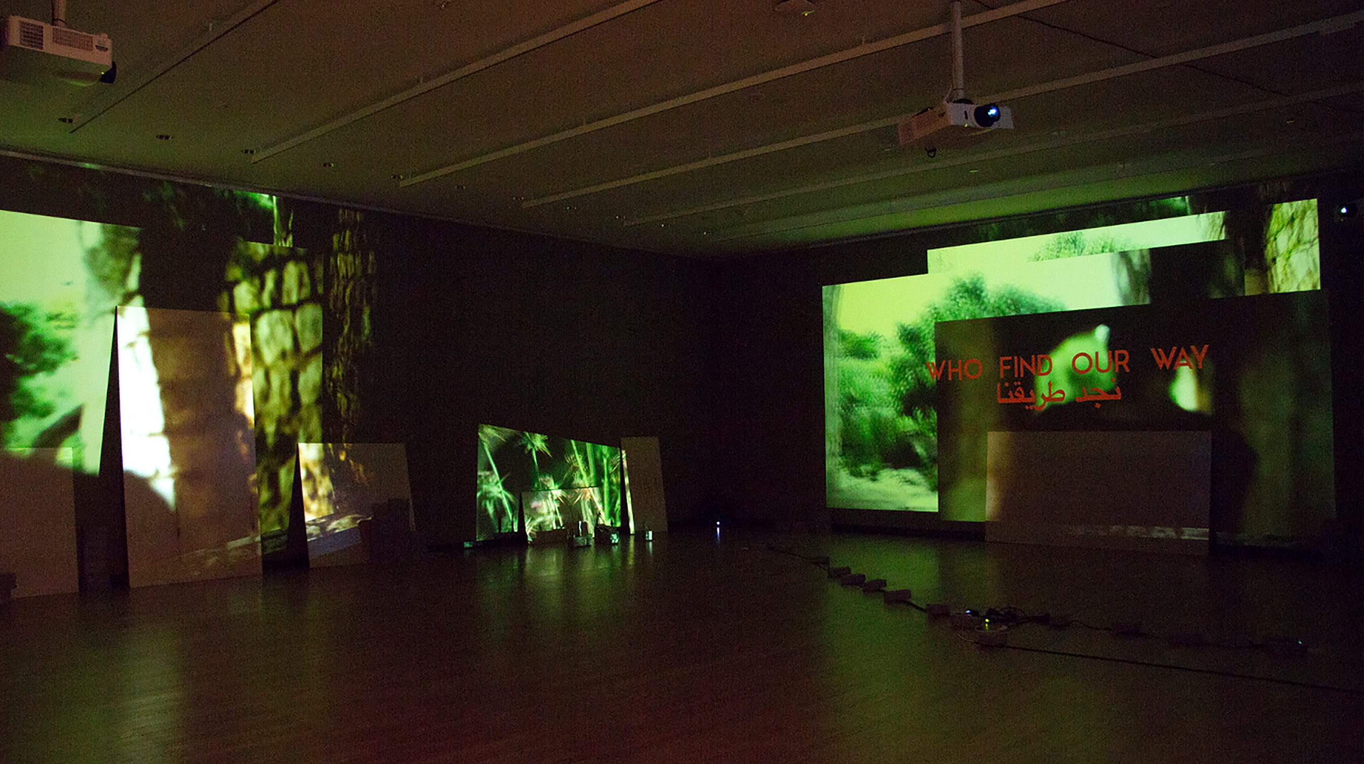 A large, empty room with video projectors hanging from the ceiling projecting images jungle langscapes on the walls. “Who Find Our Way” is written in red on one wall in English and Arabic.