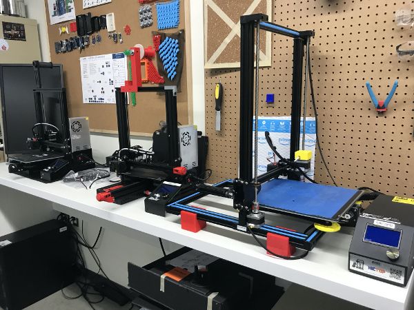 A 3D printer and related equipment and materials sit atop a work surface that is attached to a wall lined with pegboard.