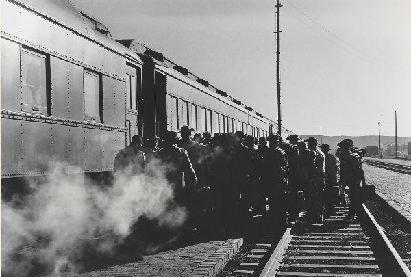 A large group of men on railroad tracks, waiting to board a train, steam in the foreground.
