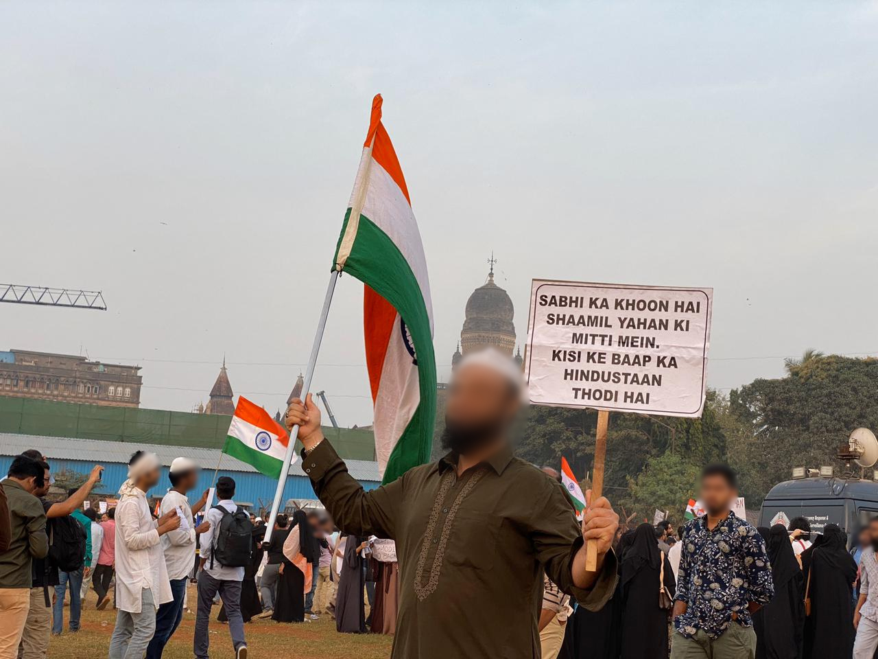 A man holds the Indian flag in one hand and a sign with poetry lines in the other. People amble in the background.