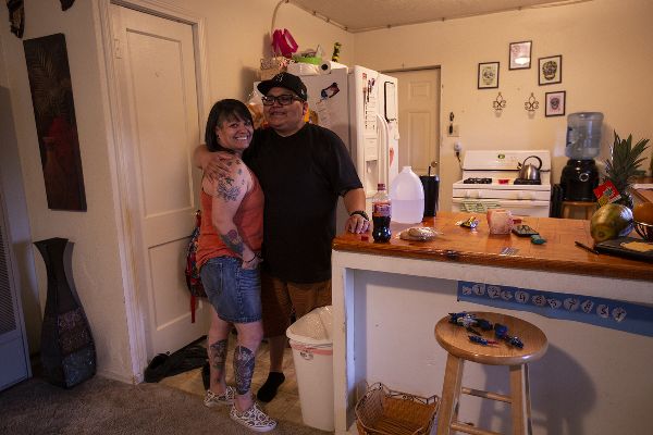 Two women embrace on a kitchen threshold, surrounded by home goods. They smile at the photographer.