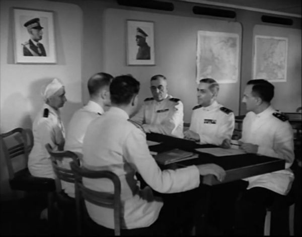 Five men in white naval uniforms and a hospital physician sit around a table. On the walls of the room are pictures of Italian political leaders.