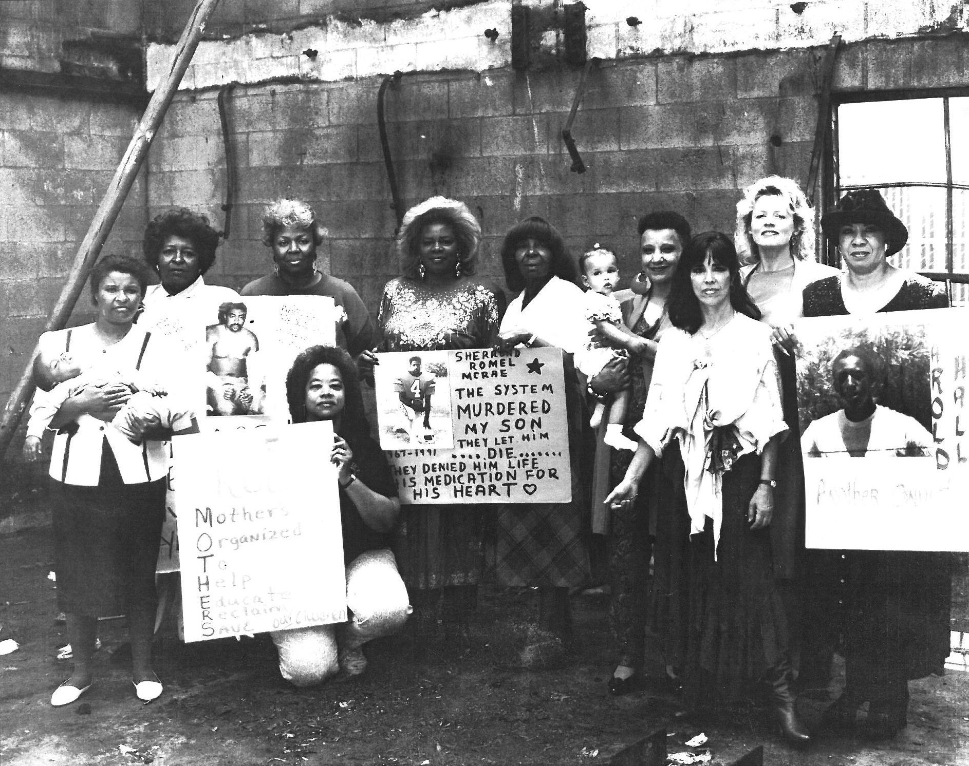 Ten women pose as a group in front of a concrete wall showing fire damage. Some hold signs and pictures, others hold small children in their arms.