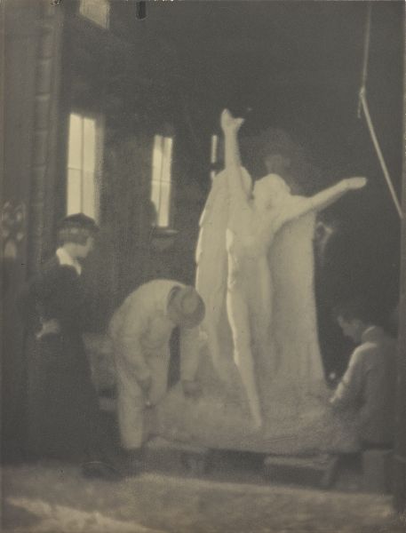 A standing woman watches two men carving the base of a marble sculpture of an angel, a third man slightly visible behind the sculpture.