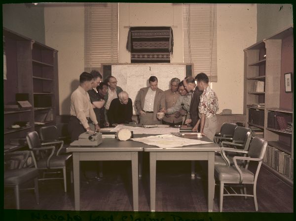 Ten men stand around the end of a conference room table, engaged in discussion.