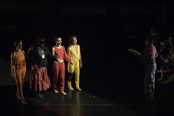 In a shaft of light, four dancers walk onto stage from the left. One wears a skirt and a dark head dress. On the right, a man stands with a hand drum.
