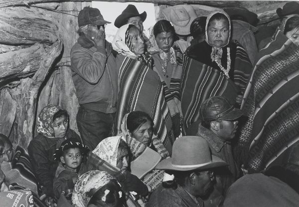 A group of people, including children, stand and sit inside a hogan. Most pay attention to activity occurring out of frame.