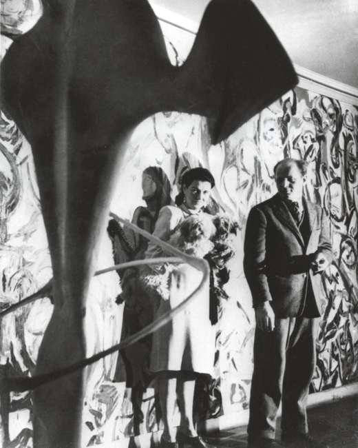 The exhibit displays Peggy Guggenheim and Jackson Pollock in her New York foyer, with Pollock in formal attire and Guggenheim holding two pet dogs, featuring Pollock’s “Mural” in the background and a sculpture by David Hare in the foreground.
