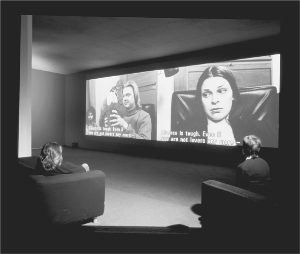 A photograph of dual screens mounted next to each other, with one showing the front view and the other shows the rear view of the same picture. Two people seated on sofas are watching the screens.